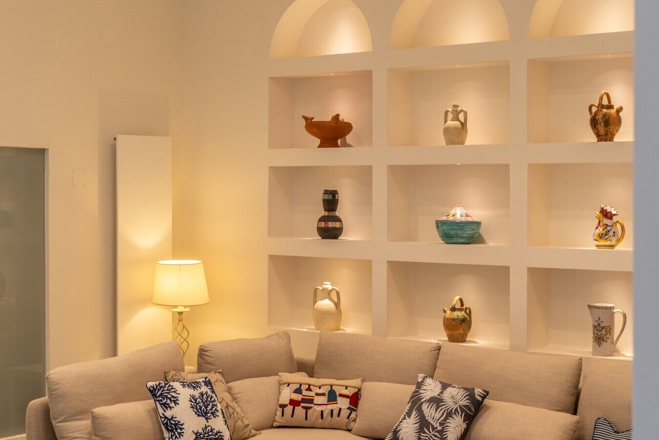Un salon comprenant un canapé, des coussins décoratifs, une lampe sur pied et un mur entier recouvert d’étagères où sont exposés divers objets.