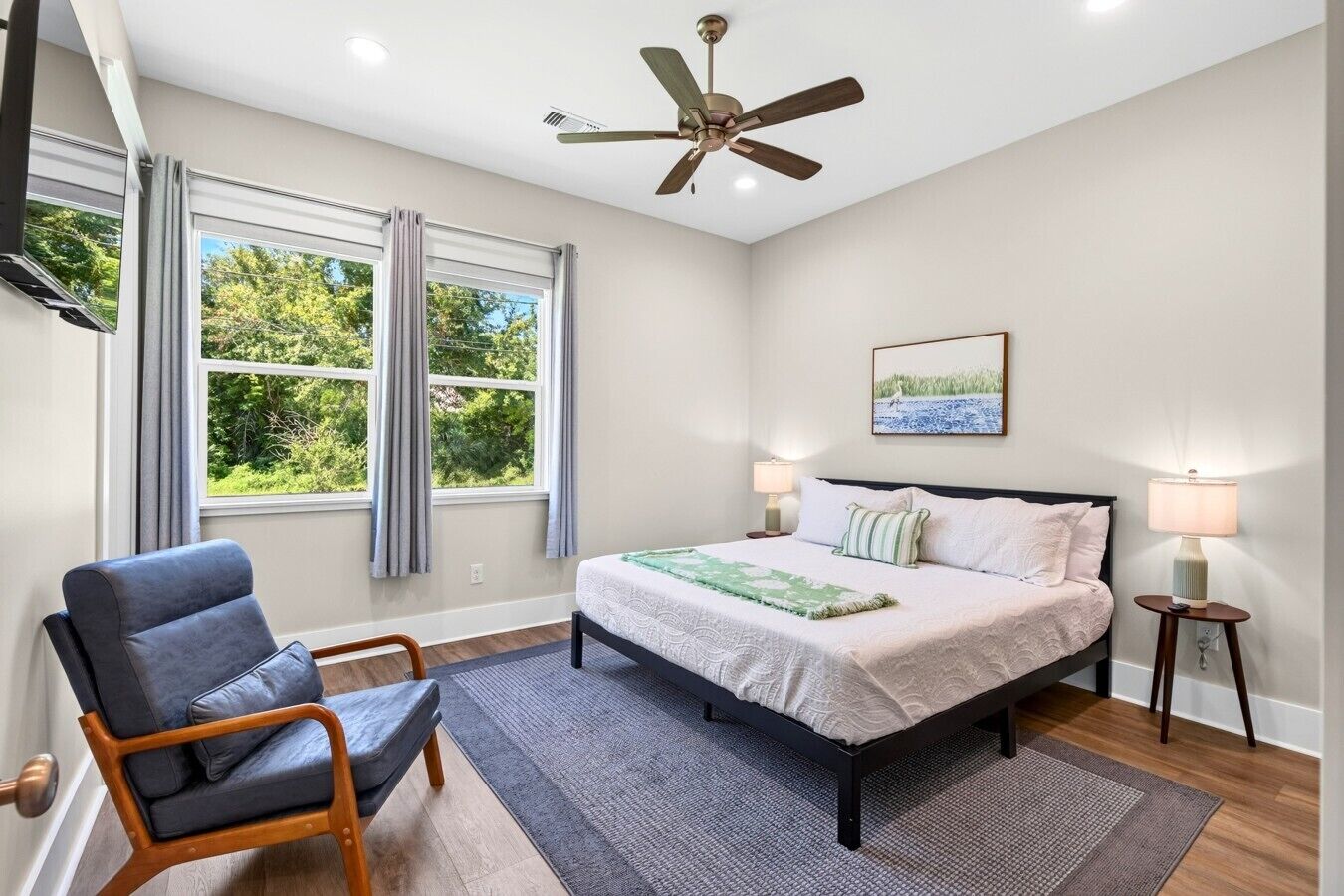 A bedroom with a bed, a chair, a ceiling fan, a window with curtains, and a nightstand with a lamp.