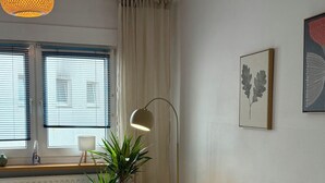 Ein Wohnzimmer mit Sofa, Stehlampe, Blumentopf, Heizkörper und einem Fenster mit Jalousien.
