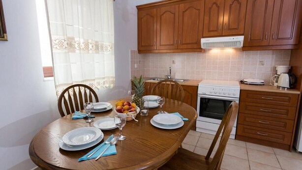 A dining area with a wooden table set for four, a kitchen with wooden cabinets and a white stove, and a window with sheer curtains. - Pleades Seaside Taygeta Gem in Mani, Near the Sea (Agios Nikolaos)