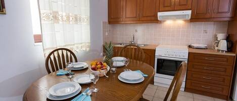 Un coin repas avec une table en bois dressée pour quatre personnes, une cuisine avec des meubles en bois et un four blanc, et une fenêtre ornée de rideaux légers.