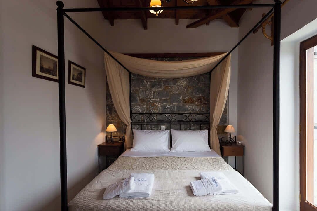 A four-poster bed with a stone wall backdrop, two bedside tables with lamps, and a window on the right.