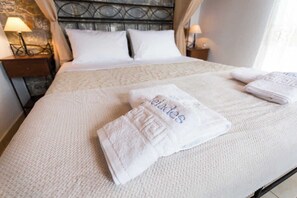 A neatly made bed with white linens and a folded towel with text, a headboard with a metal design, and bedside lamps on wooden nightstands. - Pleades Seaside Asterope Gem in Mani, Near the Sea (Agios Nikolaos)