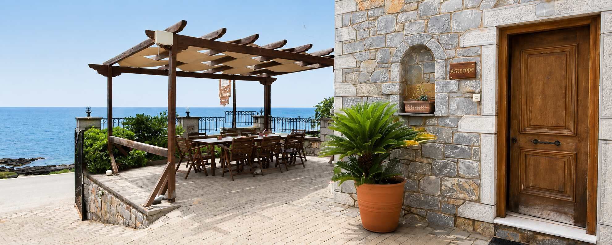 Ein Steinhaus mit einer Holztür, einer Pergola mit Tisch und Stühlen und Blick aufs Meer.