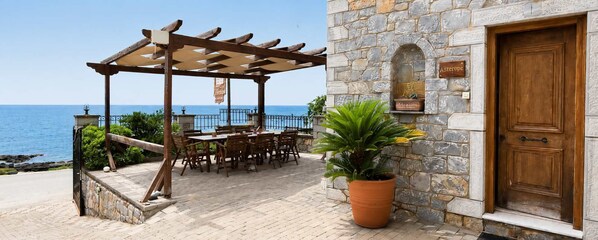 Un bâtiment en pierre avec une porte en bois, une pergola avec une table et des chaises, et une vue sur la mer.