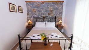 A bedroom with a stone wall, a metal bed frame, two bedside tables with lamps, a wooden table with a vase and a glass, and two framed pictures on the wall. - Pleades Seaside Alcyone Gem in Mani, Near the Sea (Agios Nikolaos)