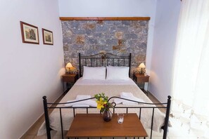 Ein Schlafzimmer mit Steinstruktur, einem Metallbettgestell, zwei Nachttischen mit Lampen, einem Holztisch mit einer Vase und einem Glas sowie zwei gerahmten Bildern an der Wand.