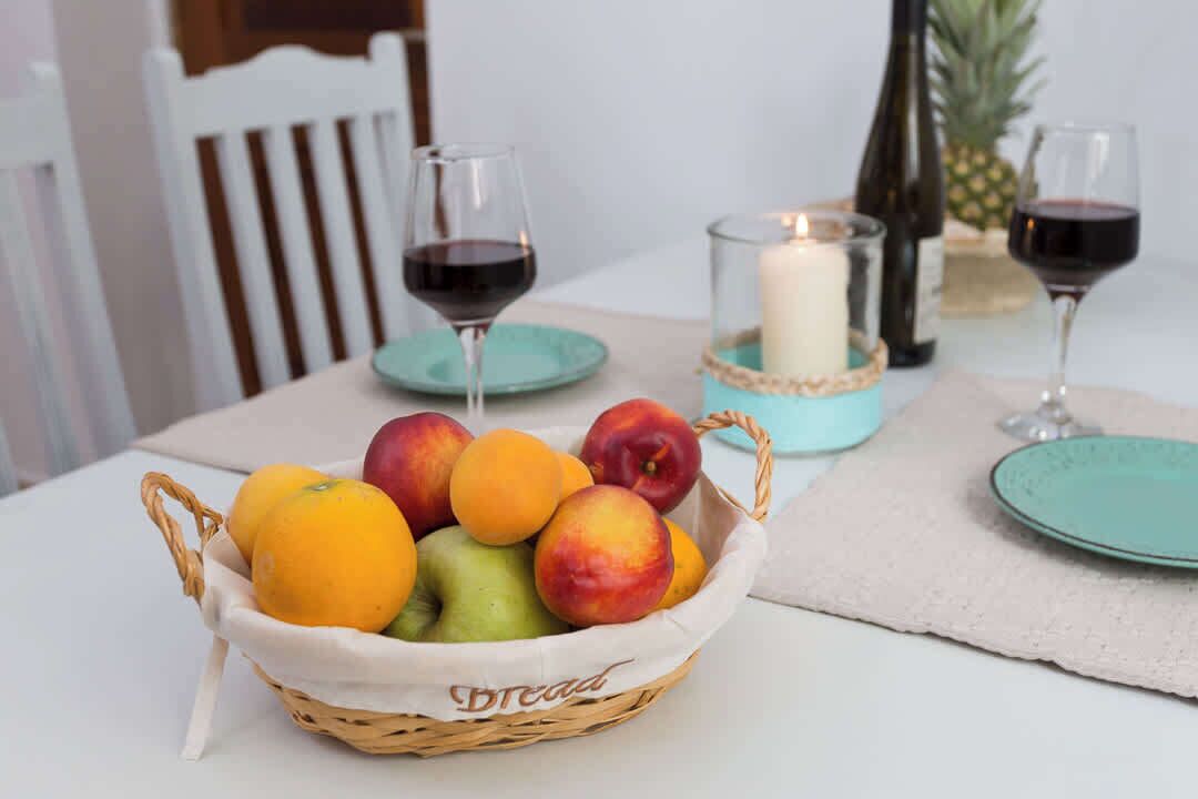 A dining table with a fruit basket, two glasses of red wine, a candle, and a pineapple.