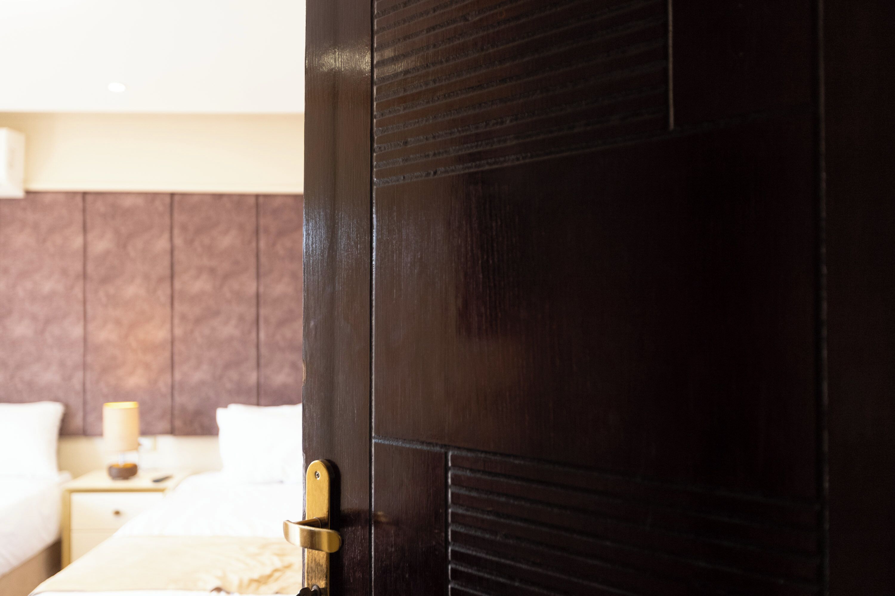 Una habitación de hotel con una puerta de madera oscura, una mesita de noche con lámpara y una cama con ropa blanca.