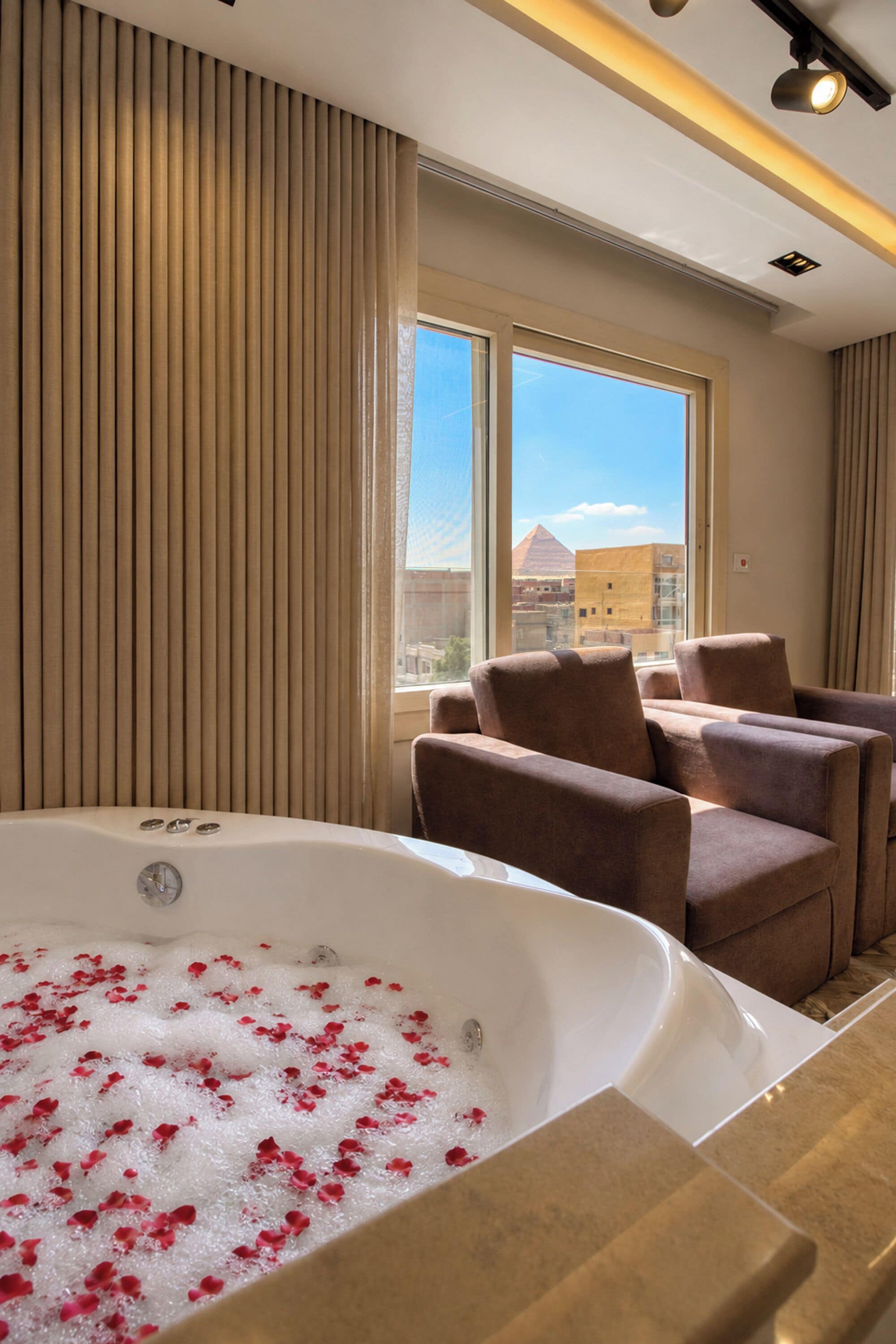 A hotel room with a large bathtub filled with bubbles and rose petals, a seating area with chairs, and a view of a pyramid through the window.