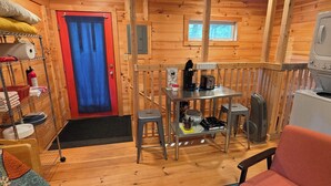 Interior de una cabaña de madera con una cama, una mesa con una silla, una pequeña cocina americana y un ventanal.