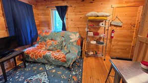 Interior acogedor de una cabaña con sofá, televisor, estante metálico con diversos objetos y alfombra estampada.
