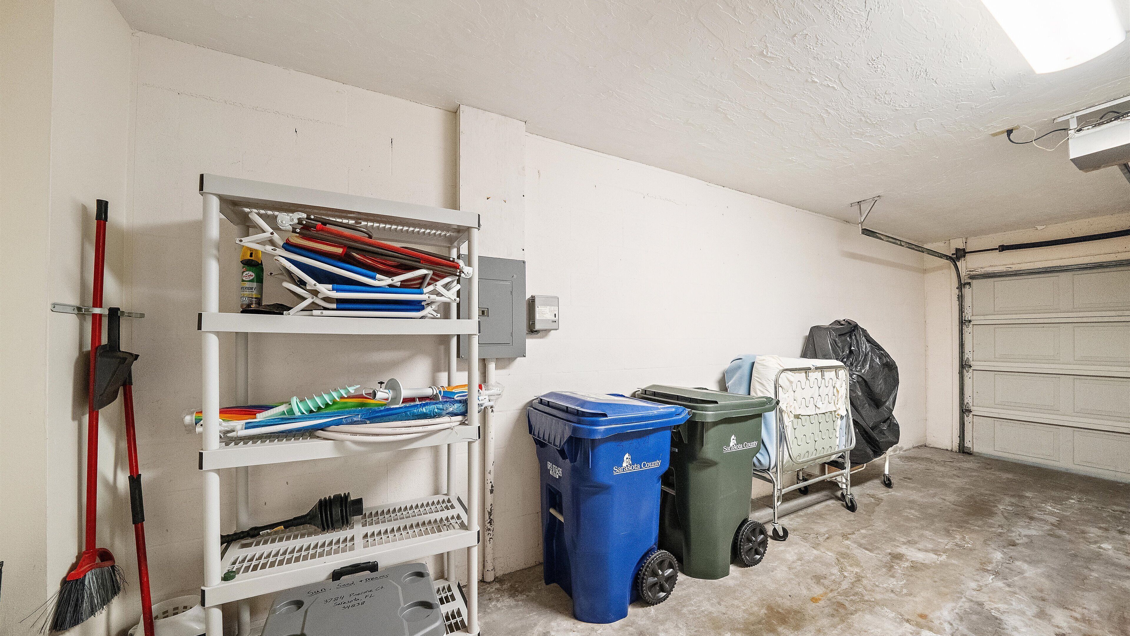 Eine Garage mit weißer Wand, einem Metallregal, einem roten Besen und zwei Müllbehältern.