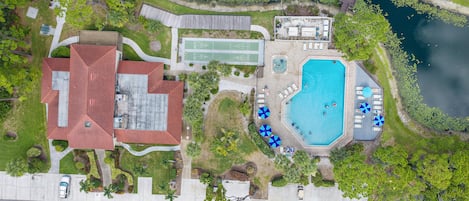 An aerial view of a property with a swimming pool, lounge chairs, a tennis court, and a covered patio area.