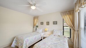 Ein Schlafzimmer mit zwei Betten, einem Deckenventilator, einer Nachttischlampe und einem Fenster mit Vorhängen.