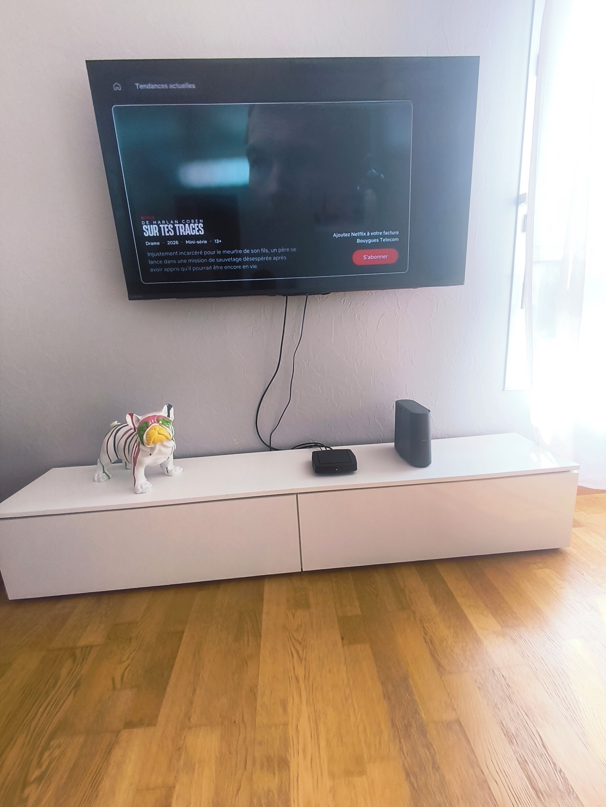 Un téléviseur à écran plat fixé au mur, un meuble blanc pour appareils électroniques en dessous, et une petite figurine posée sur le meuble.