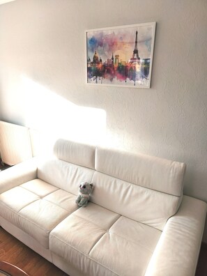 A white leather sofa with a plush cushion, a small plush toy, and a framed cityscape painting on the wall. - Comfortable apartment—5-minute walk to the RER An at Busy-Saint-Georges—8 minutes to Disney. (Bussy-Saint-Georges)