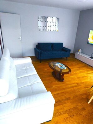 A living room with a white sofa, a blue sofa, a coffee table, and a wall-mounted TV. - Comfortable apartment—5-minute walk to the RER An at Busy-Saint-Georges—8 minutes to Disney. (Bussy-Saint-Georges)