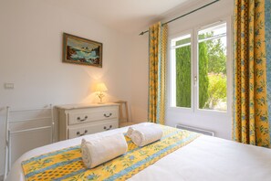 A bedroom with a bed, a bedside table with a lamp, a dresser, a painting on the wall, and a window with curtains.