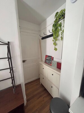 A small, white-walled room with a wooden floor, a white dresser with drawers, a black metal shelf with a hanging plant, and a black chair. - Studio overlooking a flower-filled courtyard in the heart of Montmartre (Paris)