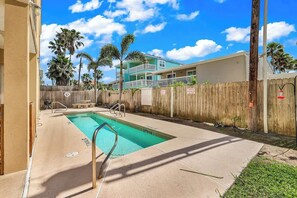 A swimming pool with a wooden fence, a palm tree, and a building in the background. - Isla Bella - 2BR Steps from the Beach with Pool (South Padre Island)