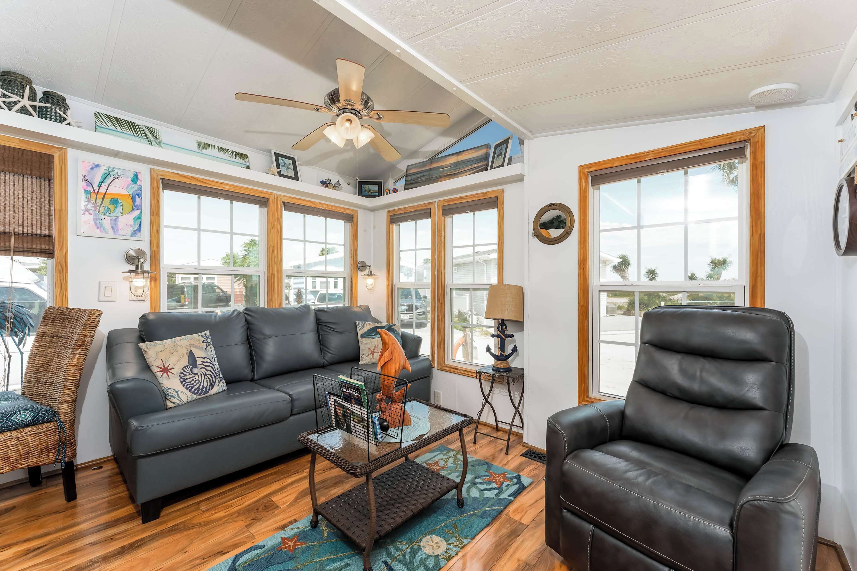 A living room with a black leather sofa, a coffee table, a wicker chair, and a leather armchair. There are large windows with wooden frames, a ceiling fan, and a view of the outside.