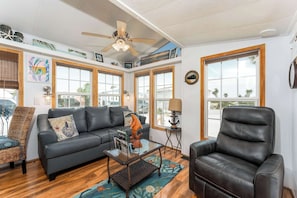 A living room with a black leather sofa, a coffee table, a wicker chair, and a leather armchair. There are large windows with wooden frames, a ceiling fan, and a view of the outside.