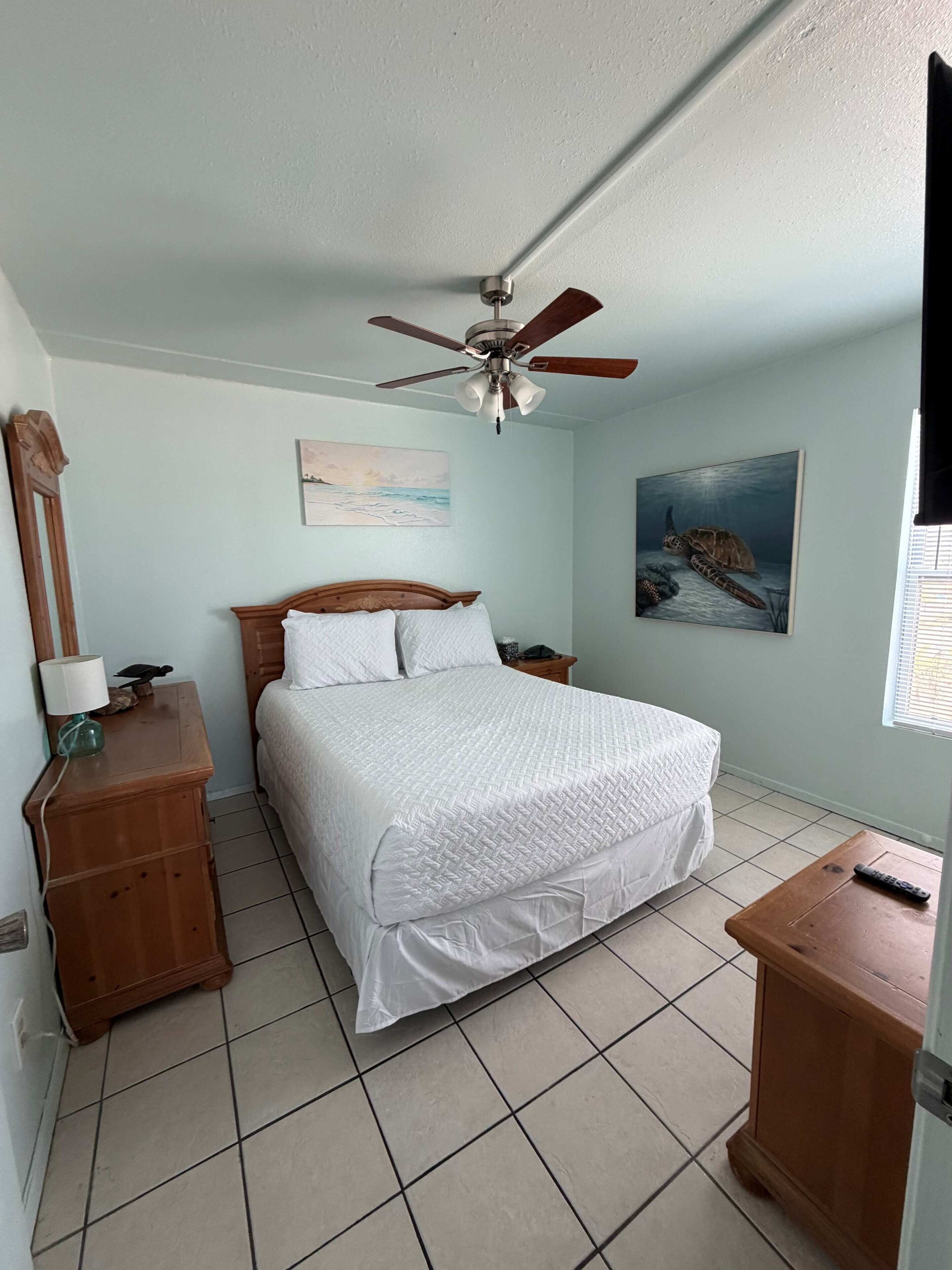 A bedroom with a bed, a ceiling fan, a nightstand, a dresser, a painting, and a window.