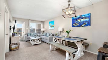 A cozy living room with a sofa, a dining table, and a bench. There are two windows with blinds, a chandelier, and artwork on the walls.