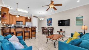 A modern living room with blue sofas, a wooden dining table, and a kitchen area with wooden cabinets and a ceiling fan. - Isla Bella - 2BR, Balcony & Pool, Beachfront Condo (South Padre Island)