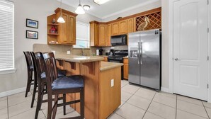 A kitchen with wooden cabinets, a stainless steel refrigerator, a microwave, and a bar area with chairs. - Isla Bella - 2BR, Balcony & Pool, Beachfront Condo (South Padre Island)
