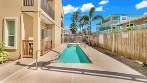 A swimming pool with a wooden fence, a building with balconies, and palm trees in the background. - Isla Bella - 2BR, Balcony & Pool, Beachfront Condo (South Padre Island)