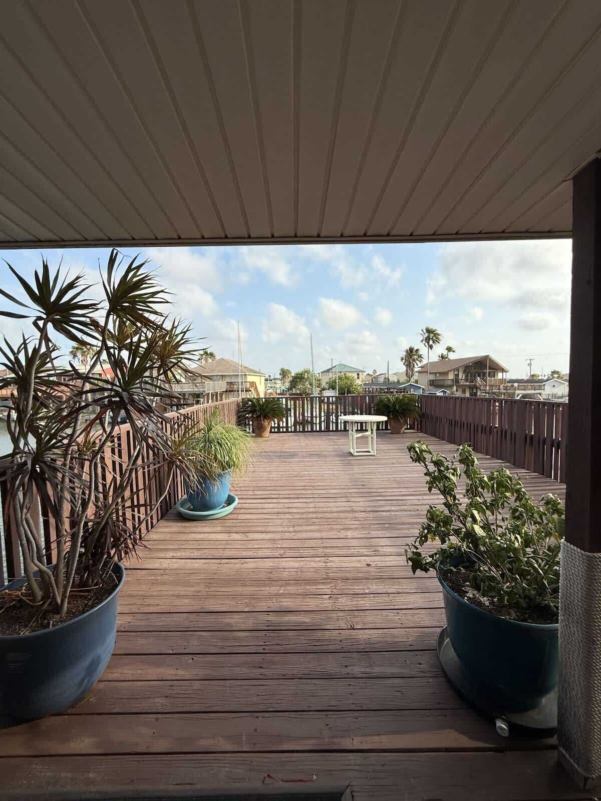 A wooden deck with potted plants on either side, leading to a view of a residential area with houses and a clear sky.