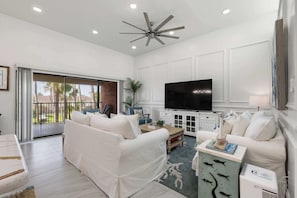 A modern living room with a ceiling fan, a flat-screen TV, white sofas, and a sliding glass door leading to a balcony. - Isla Bella - 2BR, Cielo del Sol Luxury Condo (South Padre Island)