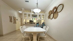 Un comedor con una mesa y sillas blancas, una cocina al fondo y platos decorativos en la pared.