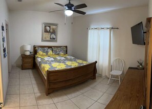 Ein Schlafzimmer mit einem Bett, einem Nachttisch, einem Sessel, einem Deckenventilator, einem Fernseher und einem Fenster mit Vorhängen.