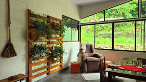 Ein Zimmer mit großem Fenster, einer Holzwand mit Pflanzen, einem Stuhl und einem Tisch mit rotem Tischläufer.