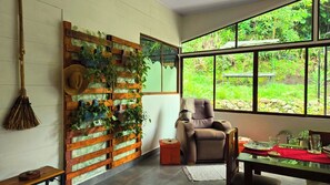 A room with a large window, a wooden wall with plants, a chair, and a table with a red tablecloth. - Charming 2-bedroom house in San Gerardo de Rivas perfect for relaxing (San Gerardo de Rivas)
