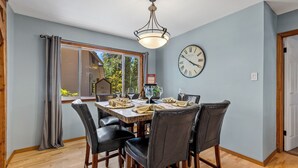 A dining area with a table set for four, a clock on the wall, and a window with curtains.