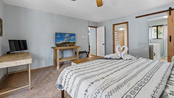 A bedroom with a bed, a desk, a television, a ceiling fan, and a door leading to another room.