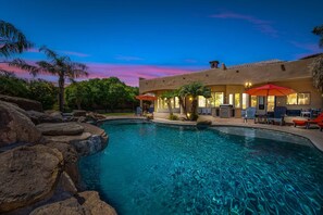 A swimming pool with rocks, palm trees, and a building with outdoor seating and umbrellas. - 104th Mesa 1331 Paradise Found (Mesa)