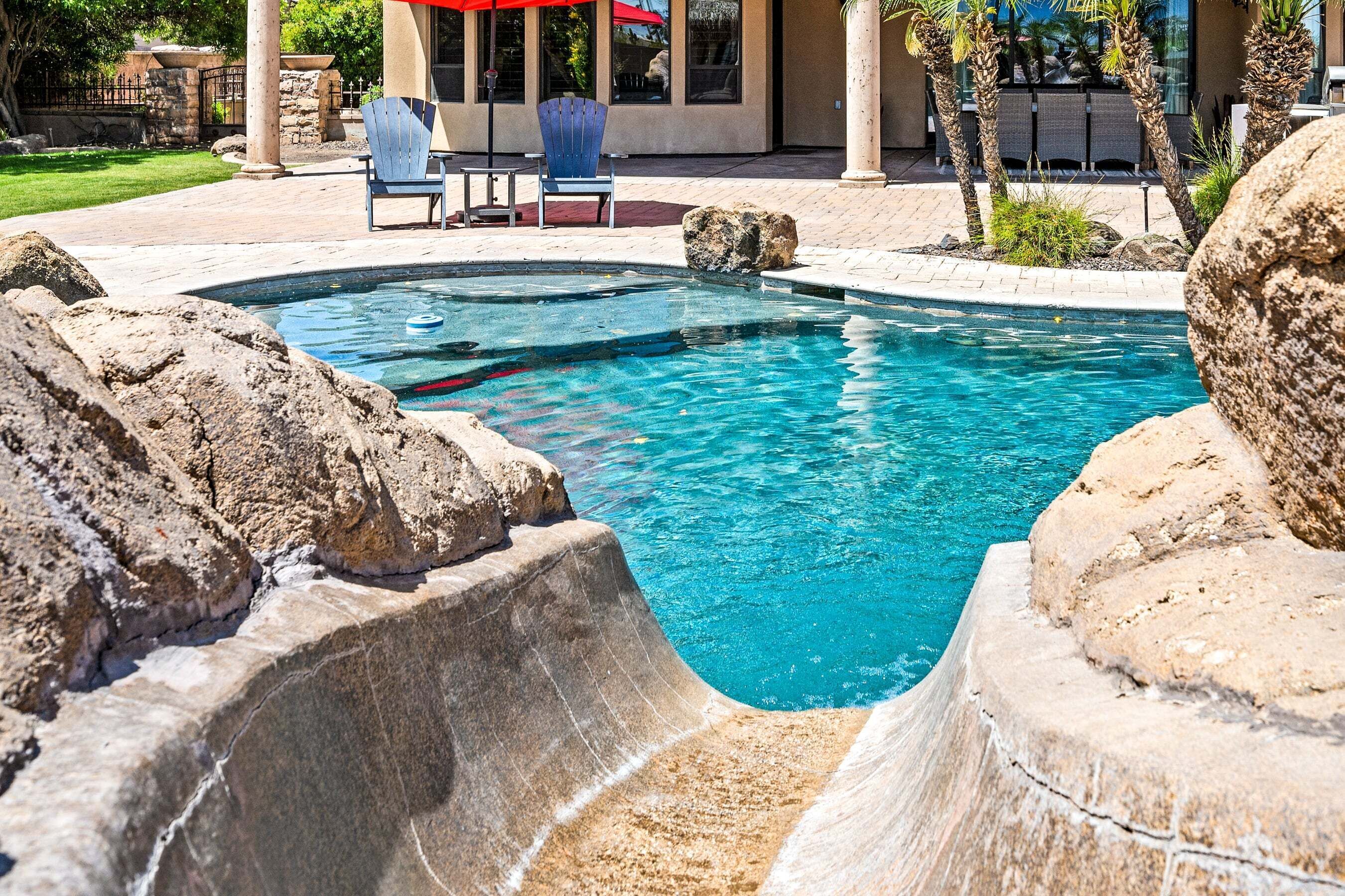 A swimming pool with a rock-lined edge, a blue chair, and a red umbrella.