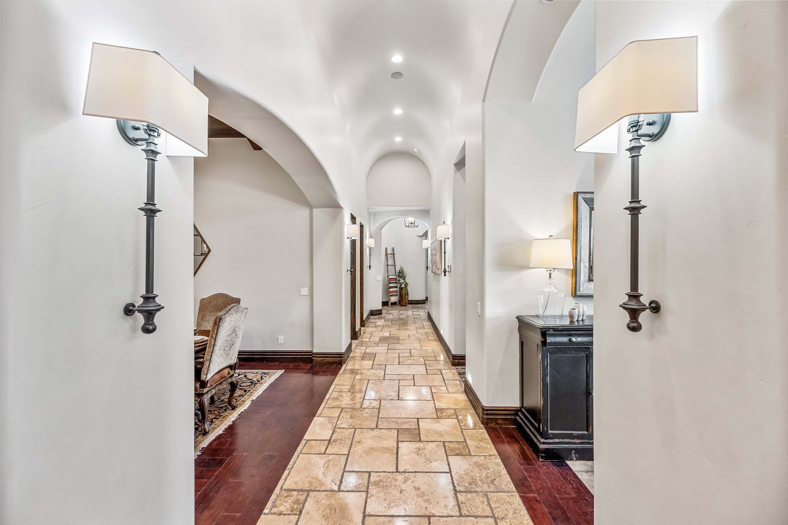 A hallway with a stone floor, wall-mounted lamps, and an arched ceiling.