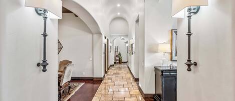 A hallway with a stone floor, wall-mounted lamps, and an arched ceiling.