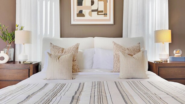 Una habitación de hotel con una cama grande, dos mesitas de noche, una obra de arte abstracta enmarcada sobre la cama y cortinas translúcidas en las ventanas.