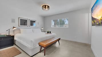A bedroom with a bed, bedside tables, a bench, a ceiling light, a window with blinds, and a wall-mounted TV.
