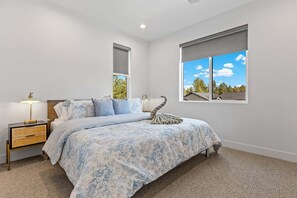A bedroom with a large bed, a nightstand, a lamp, a window with a view of trees, and a ceiling light. - Valerian Flagstaff (Flagstaff)
