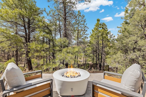 A fire pit surrounded by two wooden chairs with cushions, set in a forested outdoor area. - Valerian Flagstaff (Flagstaff)