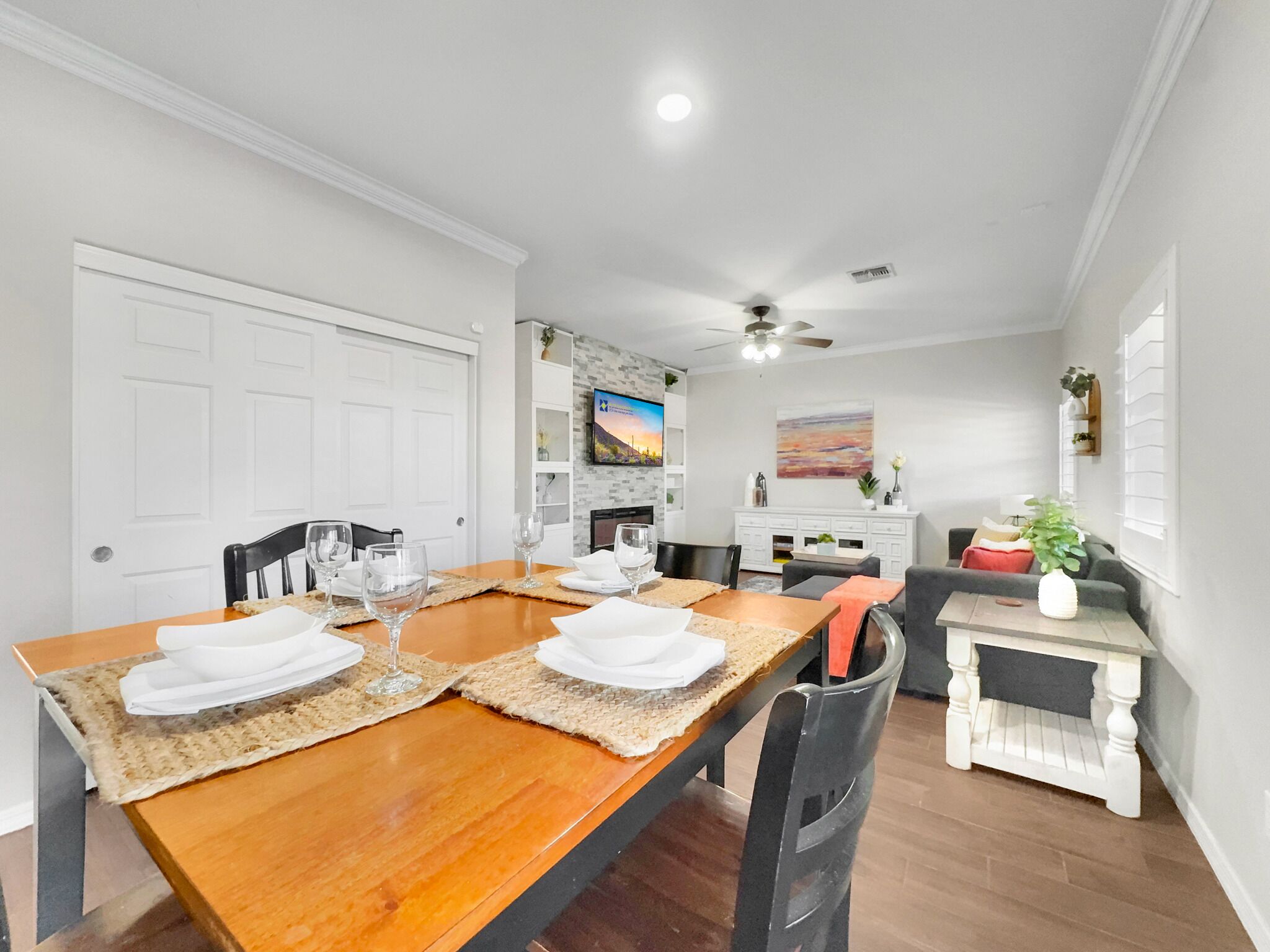 A dining area with a wooden table set for four, surrounded by chairs. There is a white cabinet with decorative items and a television mounted on the wall.