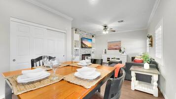 A dining area with a wooden table set for four, surrounded by chairs. There is a white cabinet with decorative items and a television mounted on the wall.